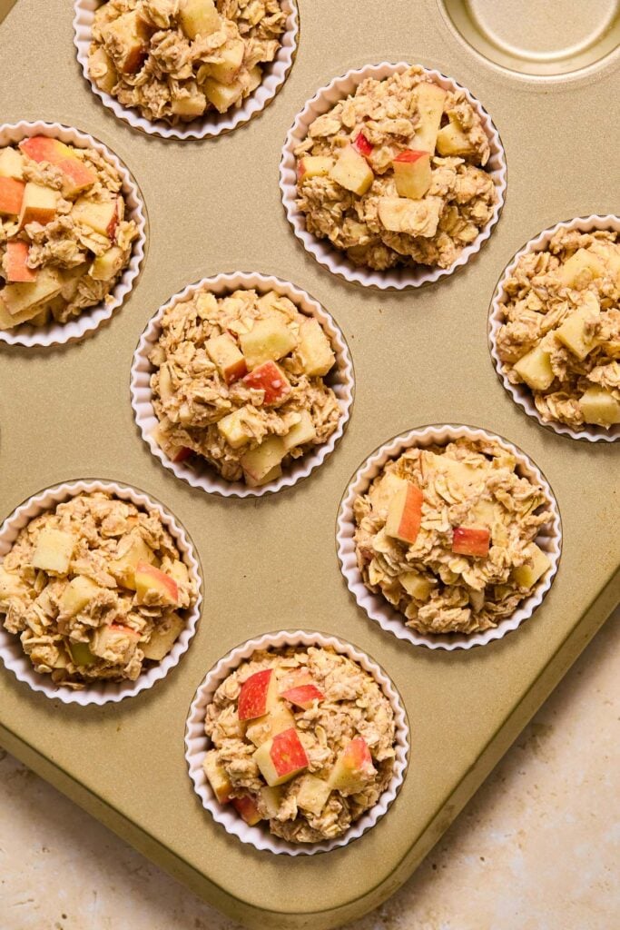 Muffin tin filled with white liners with oatmeal apple muffins in them before they are baked.