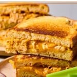 Two halves of a buffalo chicken grilled cheese stacked on top of each other on a plate with carrots and celery sticks. Behind the plate is another plate with a grilled cheese.