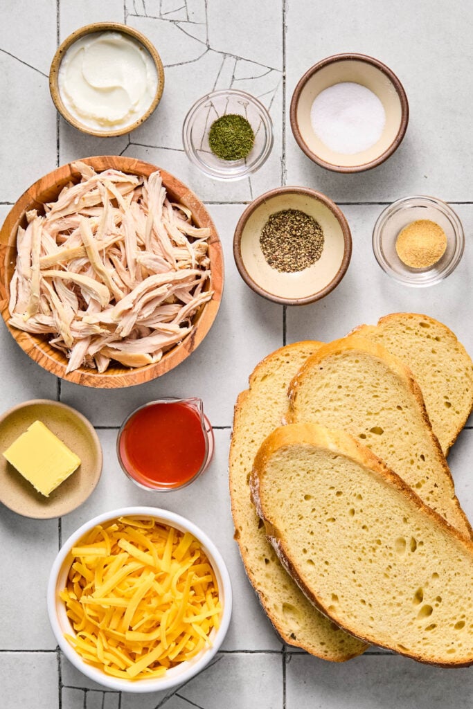 A small bowl of salt, a small bowl of dried dill, a small bowl of black pepper, a small bowl of garlic power, four slices of sourdough bread, a bowl of shredded cheddar cheese, a small bowl of buffalo sauce, a small dish with a tablespoon of butter, a bowl of shredded chicken, and a small bowl of greek yogurt on a white tile counter.