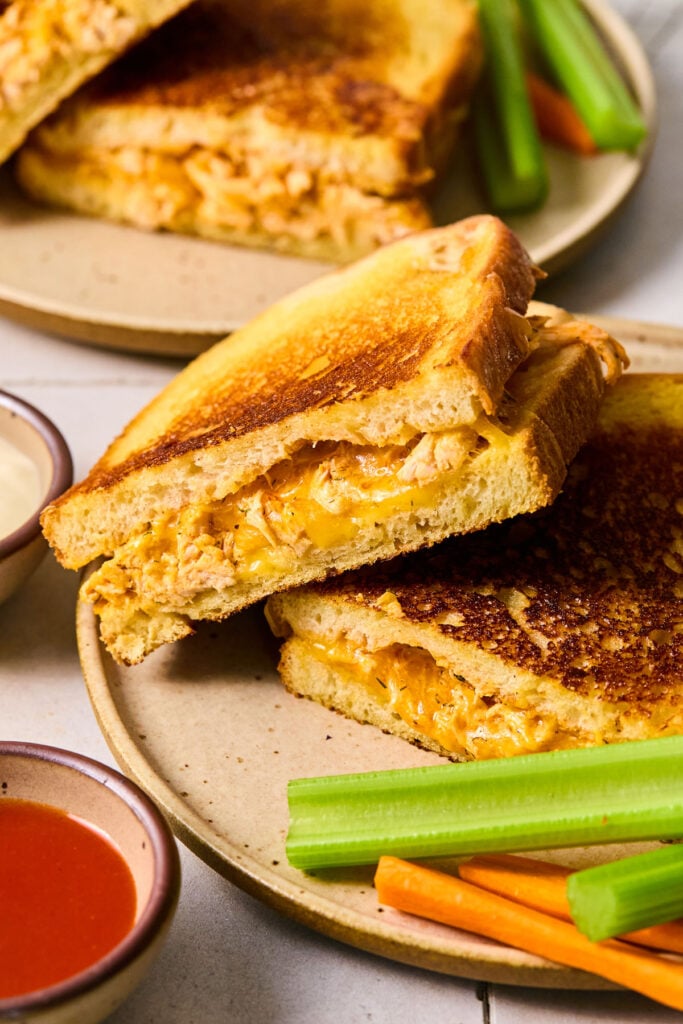 A plate with a buffalo grilled cheese cut in half and stacked on top of each other. There are celery and carrot sticks on the plate. Around the plate is a bowl of buffalo sauce and another plate with a grilled cheese and veggie sticks.