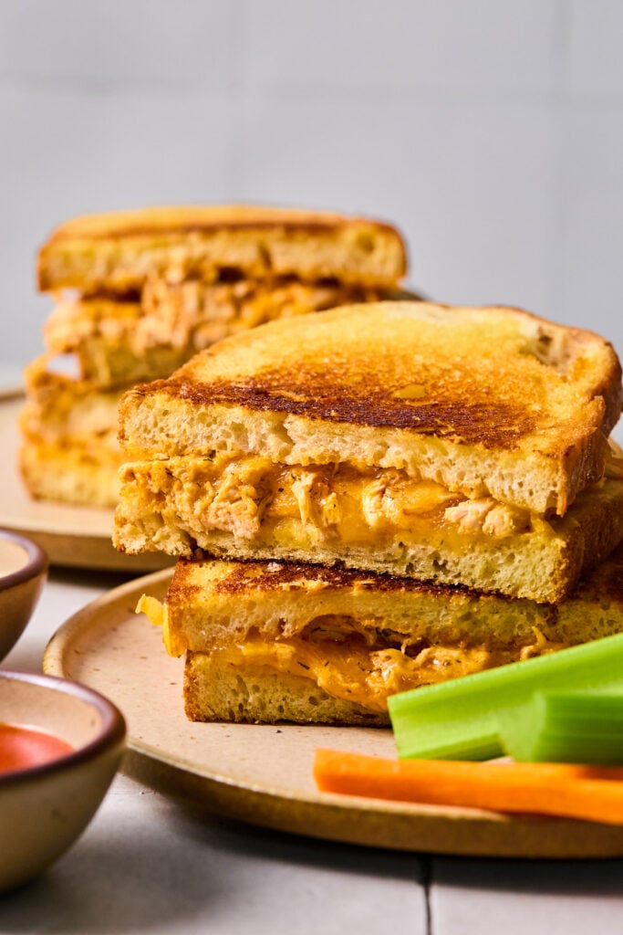 Two halves of a buffalo wing grilled cheese sandwich stacked on top of each other on a plate. Some celery and carrot sticks are also on the plate. Behind the plate is another grilled cheese stacked on top of each other, and in front of the plate is a bowl of buffalo sauce.