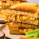 Two halves of a buffalo chicken grilled cheese stacked on top of each other on a plate with carrots and celery sticks. Behind the plate is another plate with a grilled cheese.