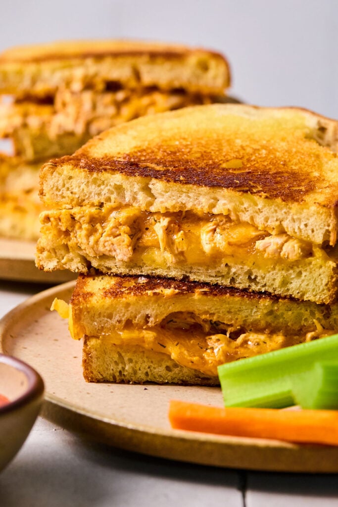 Two halves of a buffalo chicken grilled cheese stacked on top of each other on a plate with carrots and celery sticks. Behind the plate is another plate with a grilled cheese.