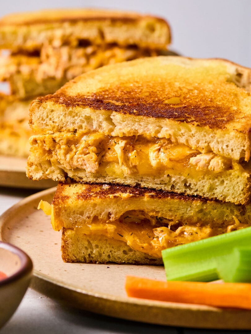 Two halves of a buffalo chicken grilled cheese stacked on top of each other on a plate with carrots and celery sticks. Behind the plate is another plate with a grilled cheese.