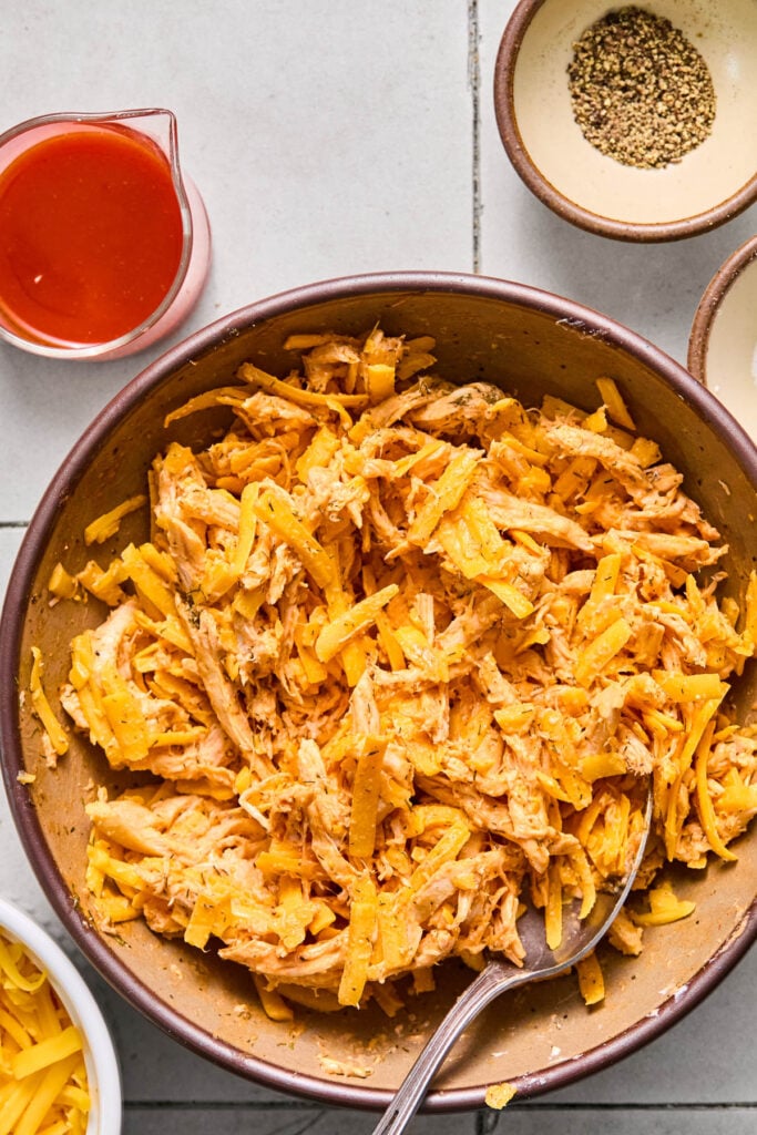 A bowl of shredded buffalo chicken mixed with shredded cheese with a spoon in it. Next to the bowl is a bowl of shredded cheese, a measuring cup of hot sauce, and a small bowl of black pepper.