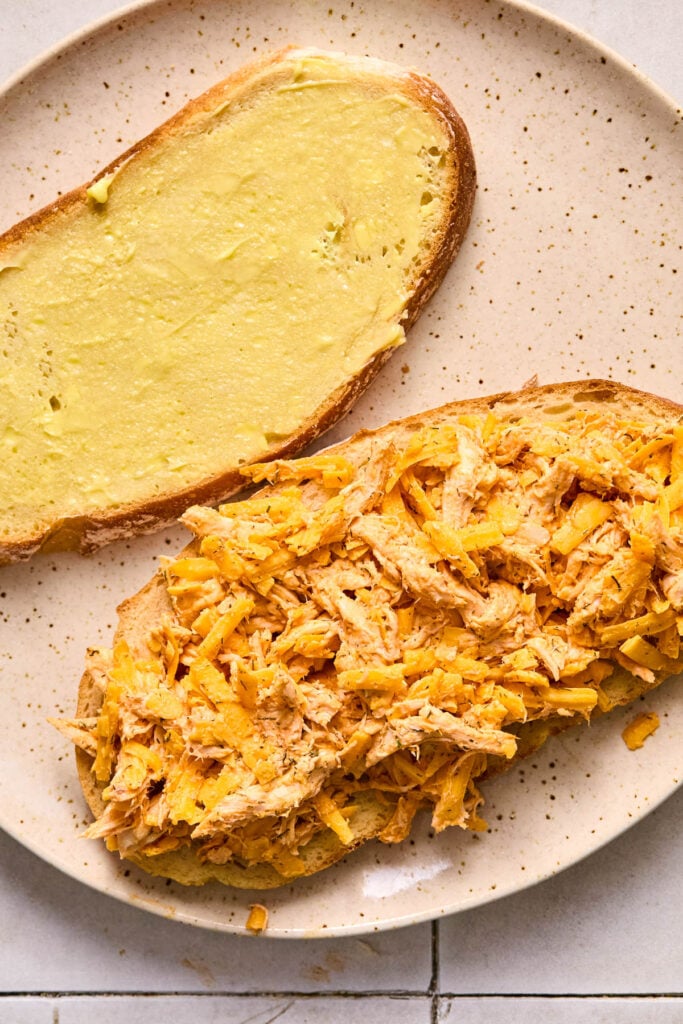 A plate with a slice of buttered bread and a slice of bread with a buffalo chicken and shredded cheese mixture on top of it.