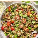 Mediterranean lentil salad with feta, seasoned chicken, veggies, olives, and fresh herbs in a large white bowl with two serving spoons. Around the bowl are two bowls of chopped herbs, a bowl of crumbled feta, some fresh herbs, and a grey kitchen towel.