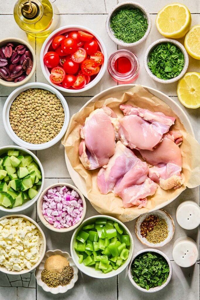 A small bowl of chopped dill, a small bowl of chopped mint, three lemon halves, a plate of raw chicken thighs, a salt and pepper shaker, a small dish of spices, a small plate of chopped parsley, a bowl of diced green bell pepper, a small dish of spices, a bowl of crumbled feta, a bowl of diced red onion, a bowl of diced cucumber, a bowl of uncooked brown lentils, a bowl of halved cherry tomatoes, a small cup of red wine vinegar, a bowl of halved kalamata olives, and an olive oil cruet on a grey tile counter.