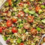 Mediterranean lentil salad with feta, seasoned chicken, veggies, olives, and fresh herbs in a large white bowl with a spoon in it. Around the bowl are some fresh herbs, a bowl of chopped herbs, a bowl of feta cheese, a bowl of sliced olives, and a blue kitchen towel.