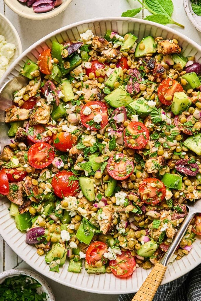 Mediterranean lentil salad with seasoned chicken, feta, veggies, olives, and fresh herbs in a large white bowl with a spoon in it. Around the bowl are some fresh herbs, a bowl of chopped herbs, a bowl of feta cheese, a bowl of sliced olives, and a blue kitchen towel.