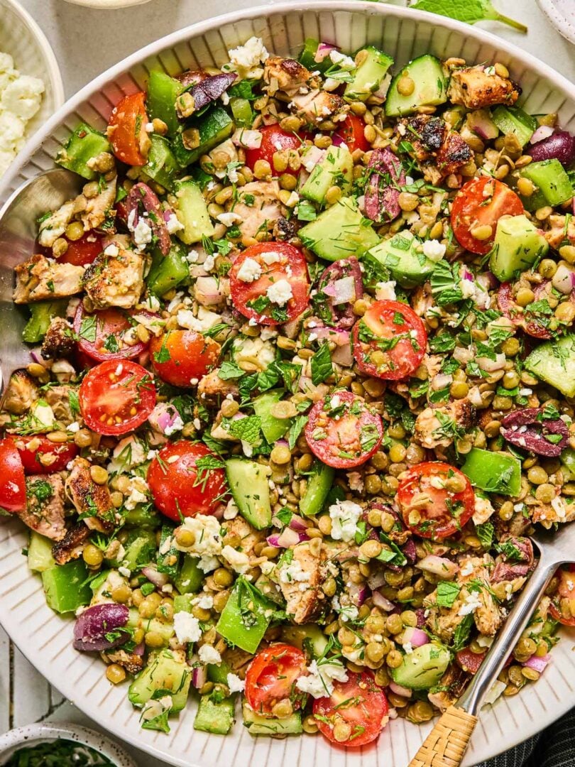 Mediterranean lentil salad with feta, seasoned chicken, veggies, olives, and fresh herbs in a large white bowl with a spoon in it. Around the bowl are some fresh herbs, a bowl of chopped herbs, a bowl of feta cheese, a bowl of sliced olives, and a blue kitchen towel.