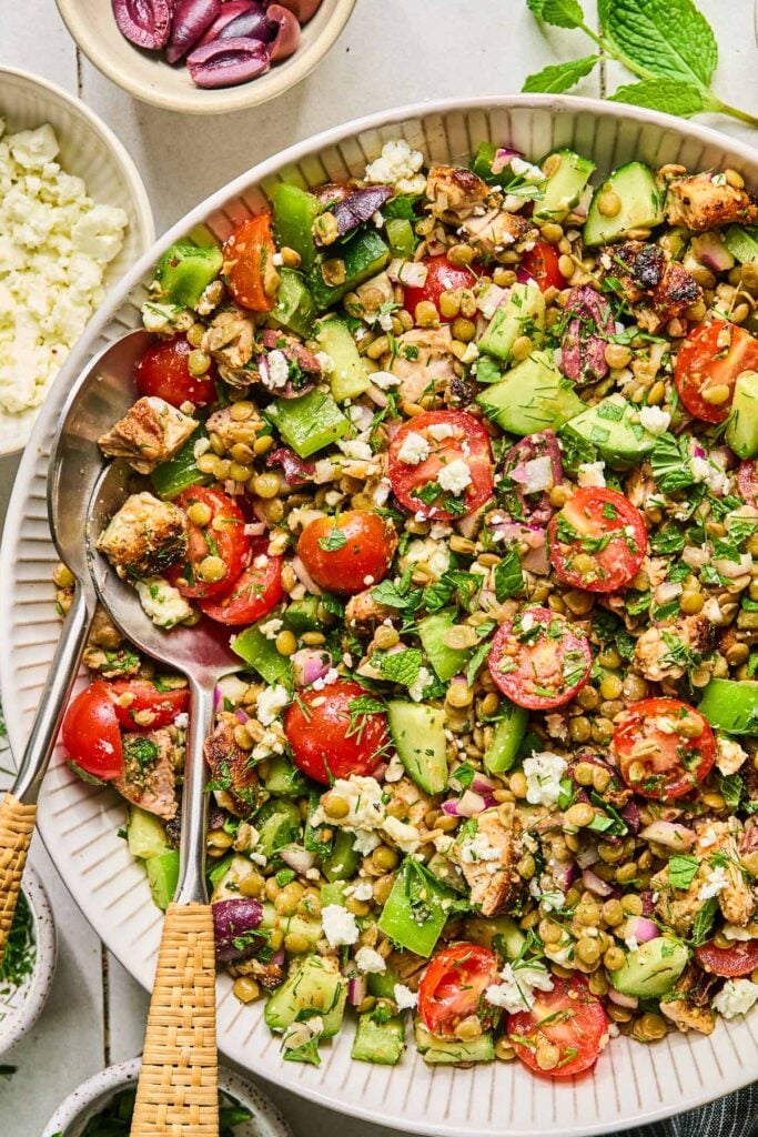 Greek lentil salad with feta, seasoned chicken, fresh veggies, olives, and herbs herbs in a white bowls with two spoons in it. Around the bowl are two small bowls of chopped herbs, a bowl of crumbled feta, a bowl of sliced olives, and some fresh herbs.