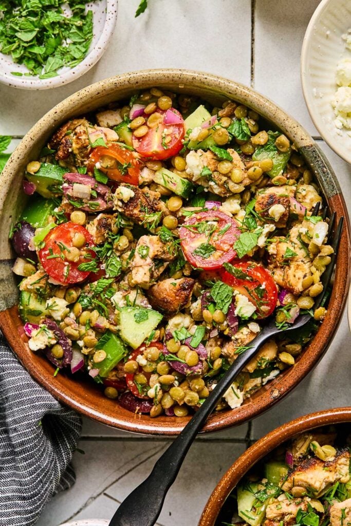 A bowl of Mediterranean lentil salad with chopped chicken, veggies, feta, olives, and fresh herbs with a fork in it. Around the bowl is another bowl of lentil salad, a blue kitchen towel, a small bowl of chopped herbs, and a bowl of crumbled feta.