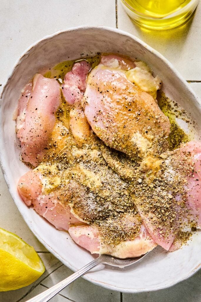 Raw chicken thighs sprinkled with spices, olive oil, and lemon juice in a baking dish with a spoon in it. Around the dish are half a juiced lemon and an olive oil cruet.
