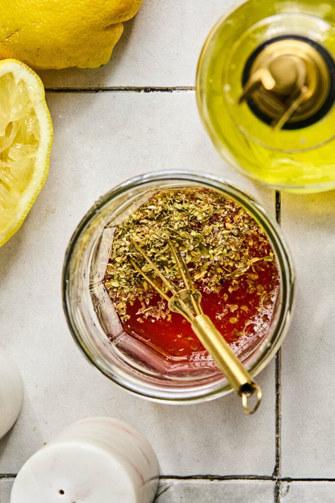 A glass jar filled with red wine vinegar, olive oil, lemon, and spices with a whisk in it. Next to it is a juiced lemon, an olive oil cruet, and a salt shaker.