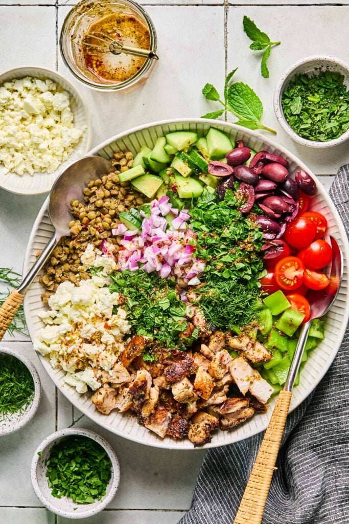 A large white bowl with cooked lentils, diced cucumber, sliced Kalamata olives, halved cherry tomatoes, diced green bell pepper, chopped seasoned chicken, crumbled feta, fresh herbs, and diced red onion with Greek vinaigrette poured overtop in it before it is mixed. Two spoons are in the bowl. Around the bowl are three small bowls of fresh herbs, a bowl of crumbled feta, an empty jar of Greek vinaigrette with a small whisk, some fresh herbs, and a grey kitchen towel.