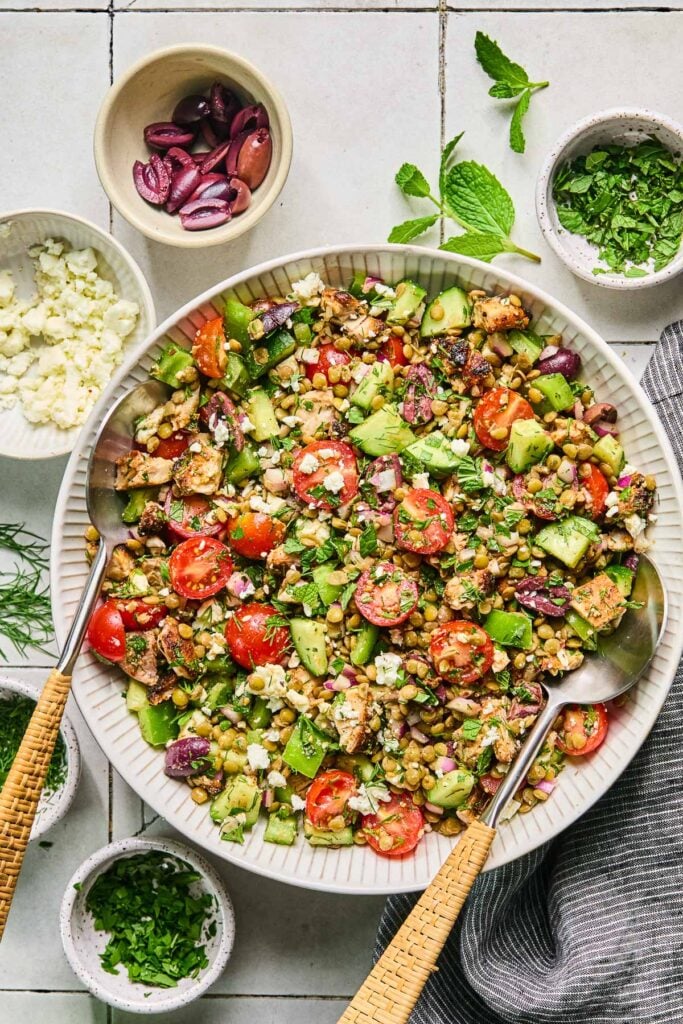 Healthy lentil salad with feta, seasoned chicken, fresh veggies, Kalamata olives, and fresh herbs tossed in a Greek vinaigrette in a white bowl with two serving spoons. Around the bowl are three bowls of chopped herbs, a bowl of crumbled feta, a bowl of sliced olives, some fresh herbs, and a grey kitchen towel.