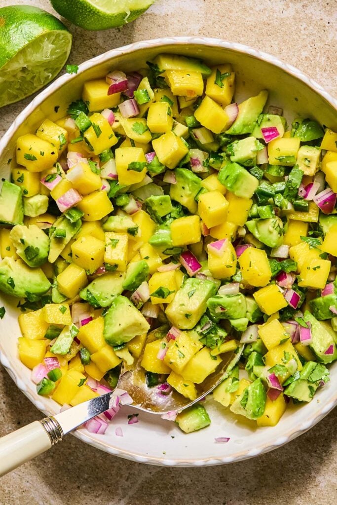 Bowl of fresh mango salsa with avocado, red onion, jalape&ntilde;o, and cilantro with a spoon in it. Around the bowl are some juiced limes.
