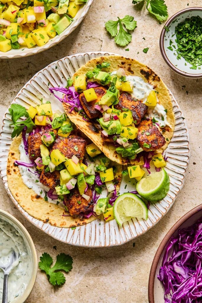 A plate of salmon tacos with mango salsa, shredded cabbage, and fish taco sauce on corn tortillas. Also on the plate are some lime wedges and cilantro. Around the plate are a bowl of shredded cabbage, a bowl of fish taco sauce, a bowl of mango salsa, and a bowl of chopped cilantro.