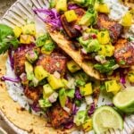 Salmon tacos on a plate with shredded cabbage, mango avocado salsa, and fish taco sauce. On the plate are some lime wedges. Around the plate are a bowl of fish taco sauce and a bowl of mango salsa.