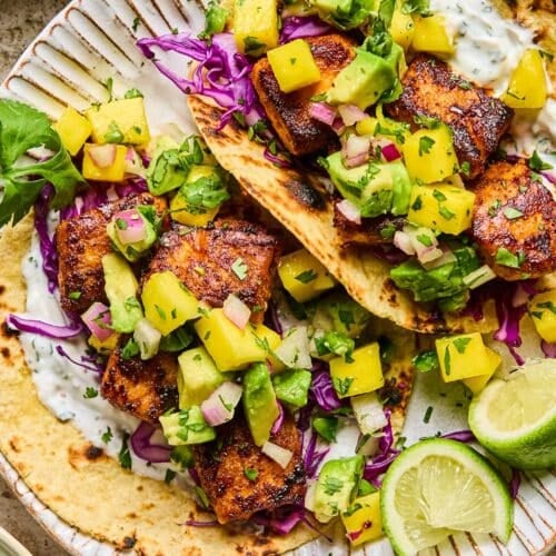 Salmon tacos on a plate with shredded cabbage, mango avocado salsa, and fish taco sauce. On the plate are some lime wedges. Around the plate are a bowl of fish taco sauce and a bowl of mango salsa.