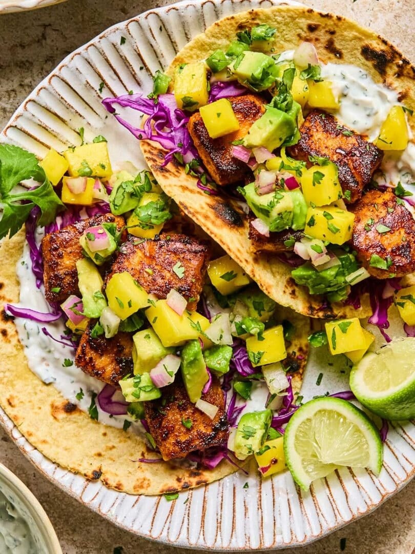 Salmon tacos on a plate with shredded cabbage, mango avocado salsa, and fish taco sauce. On the plate are some lime wedges. Around the plate are a bowl of fish taco sauce and a bowl of mango salsa.