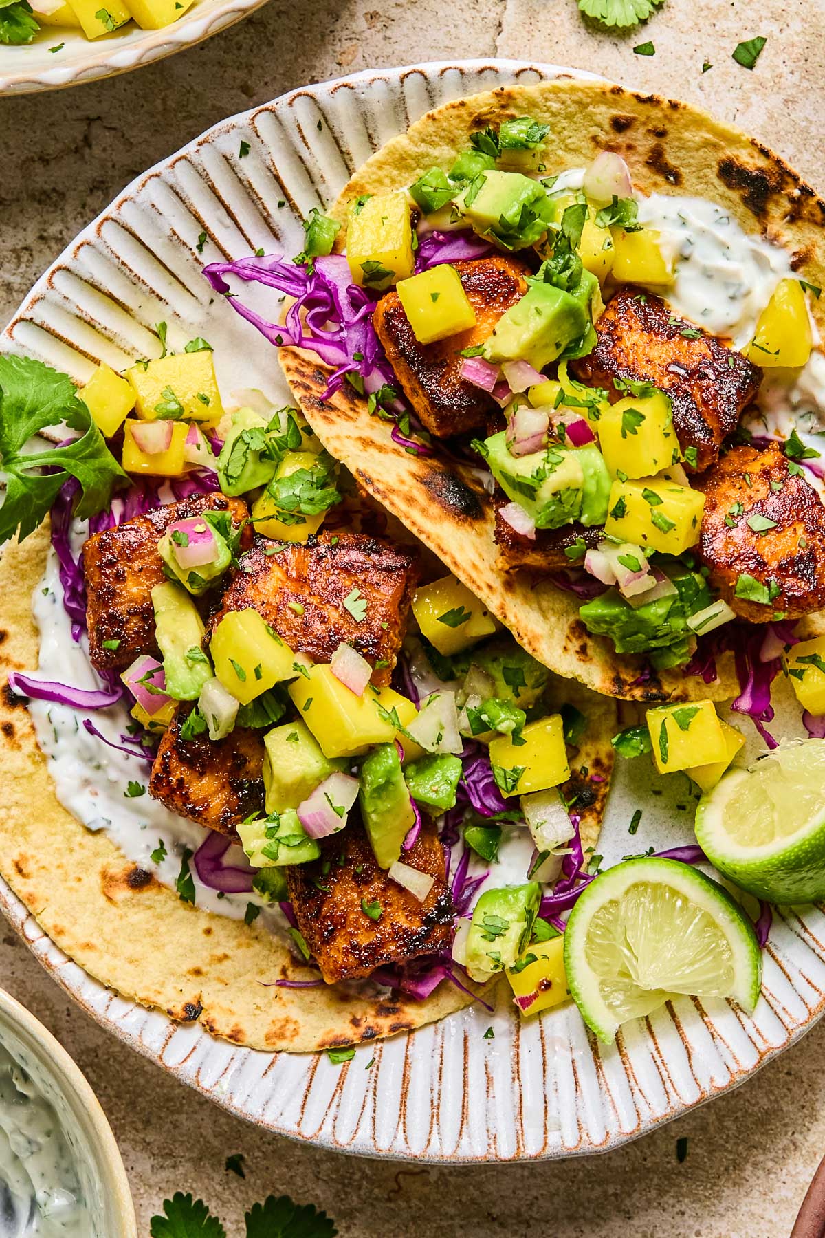 Salmon tacos on a plate with shredded cabbage, mango avocado salsa, and fish taco sauce. On the plate are some lime wedges. Around the plate are a bowl of fish taco sauce and a bowl of mango salsa.