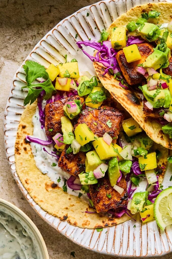 A close-up of salmon fish tacos with fish taco sauce, red cabbage, and mango avocado salsa on a plate with lime wedges. Next to the plate is a bowl of fish taco sauce.