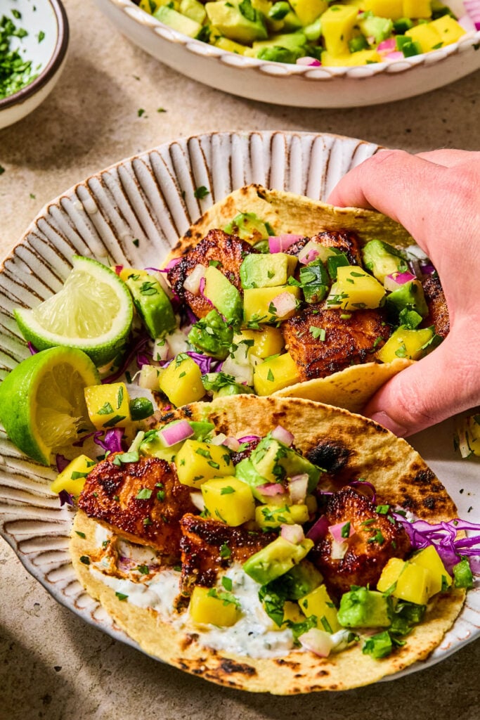 A plate of blackened salmon tacos with mango salsa, cabbage, and fish taco sauce, and lime wedges. A hand is picking up one of the tacos. Behind the plate is a small bowl of chopped cilantro and a bowl of mango salsa.