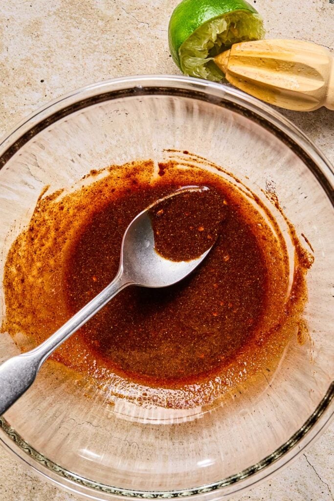 Bowl filled with a brownish red marinade with a metal spoon in it. Next to it is half a lime and a wooden reamer.