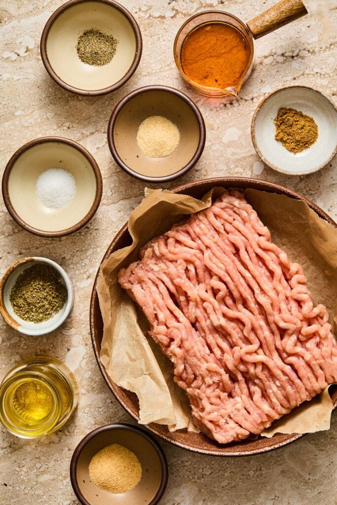 A measuring cup of maple syrup, a small bowl of cumin, a plate with a pound of raw ground chicken, a small bowl of garlic powder, a small jar of olive oil, a small bowl of dried sage, a small bowl of salt, a small bowl of onion powder, and a small bowl of black pepper on a tan concrete counter.