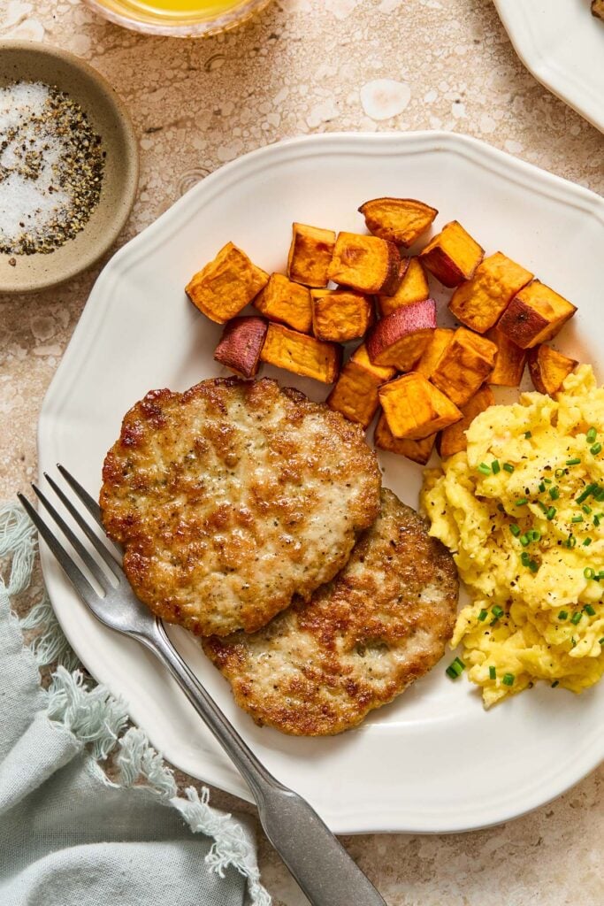 A plate of diced roasted sweet potatoes, scrambled eggs, and two chicken maple breakfast sausage patties with a fork on it. Around the place are a green napkin and a small dish of salt and pepper.