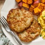 A close-up of a plate with two chicken breakfast sausage patties, roasted sweet potatoes, and scrambled eggs with a fork on it. Next to the plate is a green napkin and a small dish of salt and pepper.