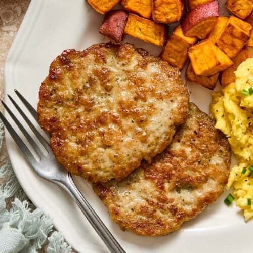 A close-up of a plate with two chicken breakfast sausage patties, roasted sweet potatoes, and scrambled eggs with a fork on it. Next to the plate is a green napkin and a small dish of salt and pepper.