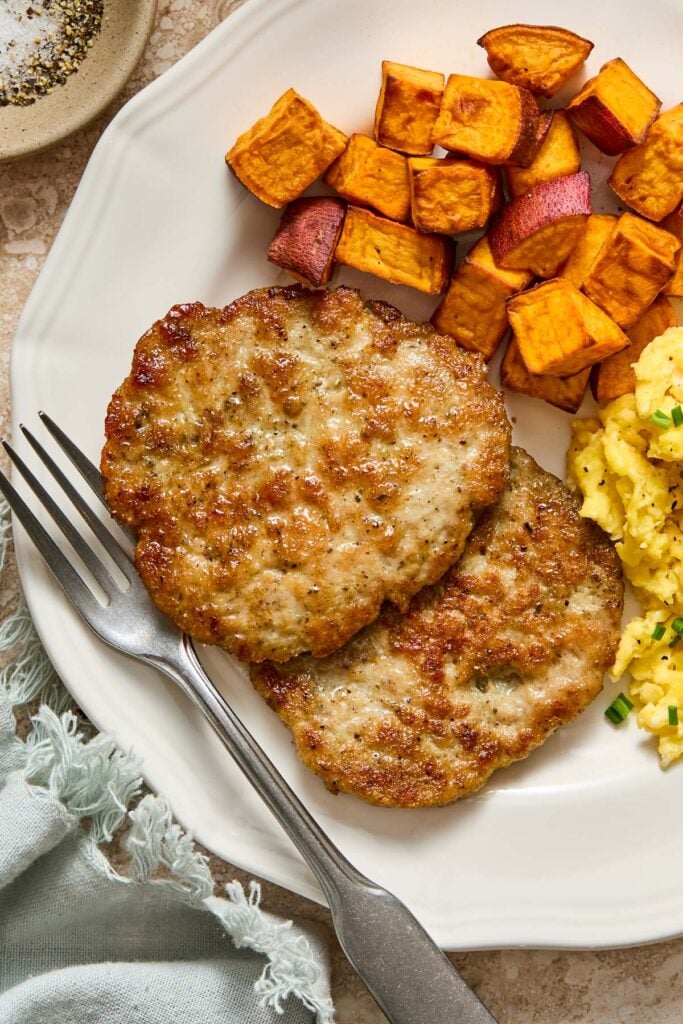 A close-up of a plate with two chicken breakfast sausage patties, roasted sweet potatoes, and scrambled eggs with a fork on it. Next to the plate is a green napkin and a small dish of salt and pepper.