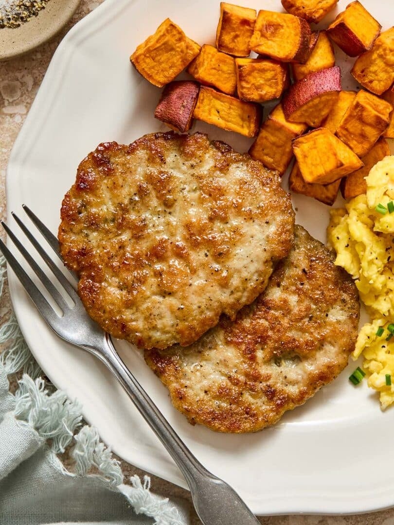 A close-up of a plate with two chicken breakfast sausage patties, roasted sweet potatoes, and scrambled eggs with a fork on it. Next to the plate is a green napkin and a small dish of salt and pepper.