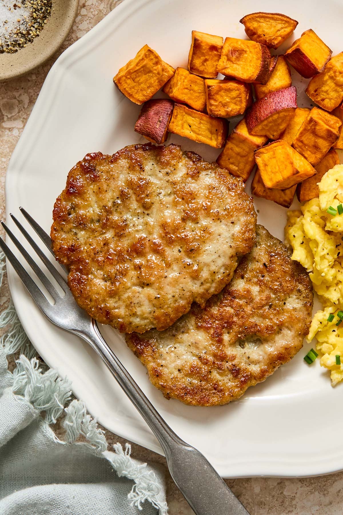 A close-up of a plate with two chicken breakfast sausage patties, roasted sweet potatoes, and scrambled eggs with a fork on it. Next to the plate is a green napkin and a small dish of salt and pepper.