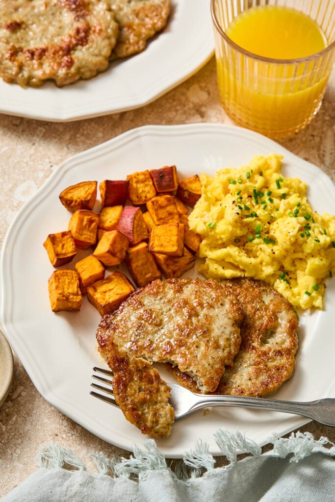 A plate with diced roasted sweet potatoes, scrambled eggs, and two chicken breakfast sausage patties. A fork is also on the plate with a piece of chicken sausage on it. Around the plate is a green napkin, a plate of more sausages, and a glass of orange juice.