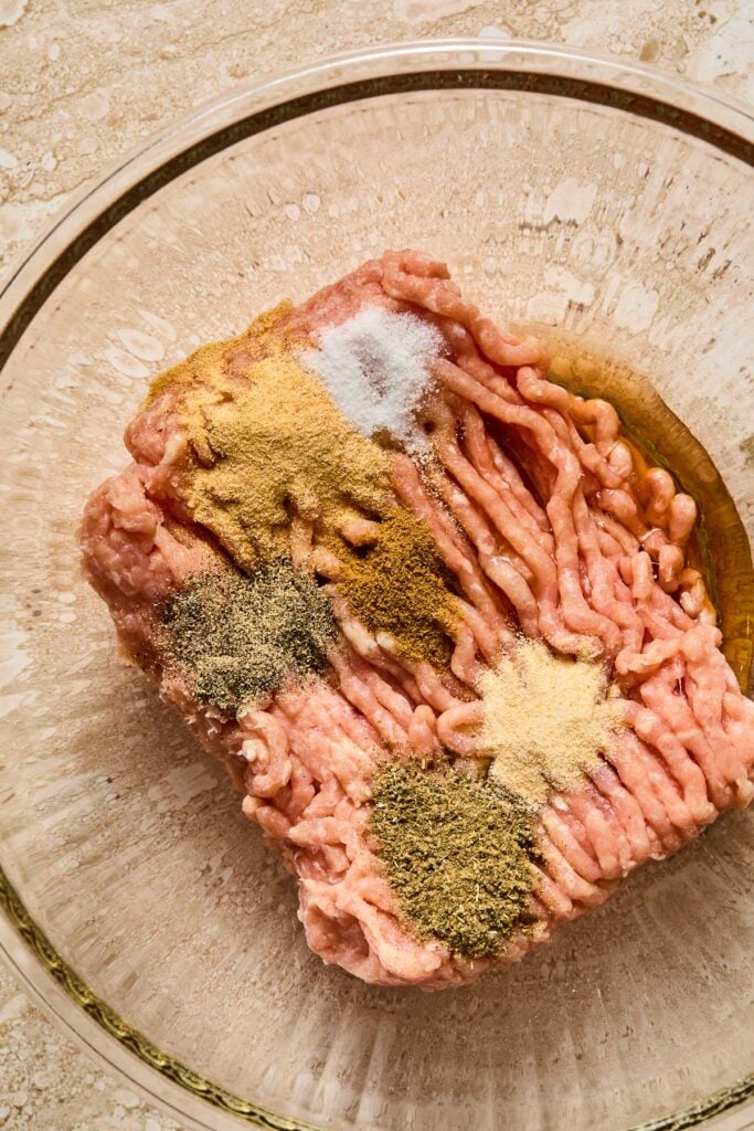 A glass mixing bowl of raw ground chicken, spices, and maple syrup before it is mixed.