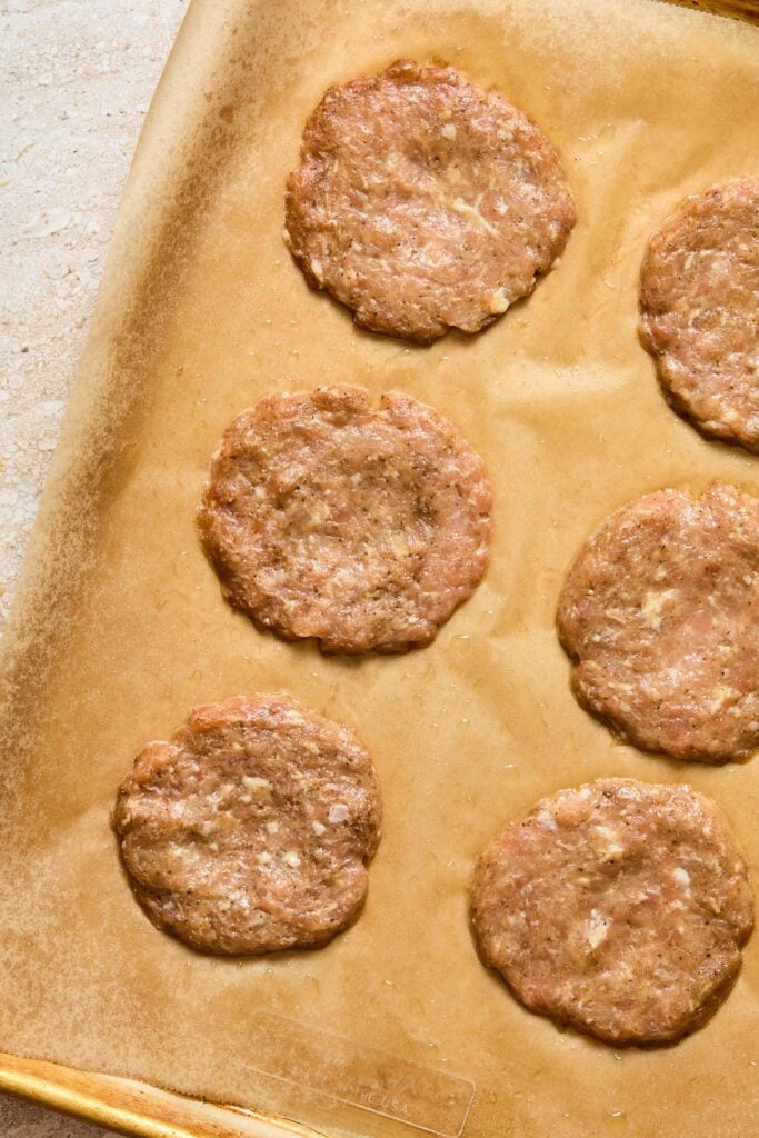 Raw ground chicken breakfast sausage patties spread out on a baking sheet lined with parchment paper.