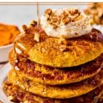 A stack of pumpkin pancakes topped with greek yogurt, pecan pieces, cinnamon, and maple syrup on a plate. Around the plate are some cinnamon sticks, pecan pieces, a bowl of pumpkin puree, and a bowl of pecans.