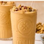 Pumpkin protein shake in a glass topped with chopped pecans and cinnamon, with a straw in it. Around the glass are some chopped pecans, a bowl of pumpkin puree, a bowl of chopped pecans, a bowl of granola, and another glass with a smoothie.