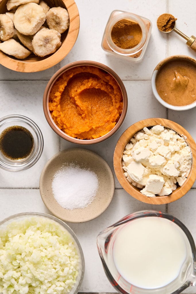 A spice jar with pumpkin pie spice, a teaspoon filled with pumpkin pie spice, a small bowl of almond butter, a bowl of pumpkin puree, a bowl of vanilla protein powder, a measuring cup of milk, a bowl of frozen cauliflower rice, a small bowl of salt, a small bowl of vanilla bean paste, and a bowl of frozen banana slices on a white tile counter.