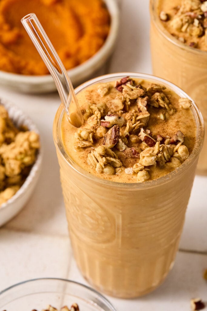 Pumpkin smoothie topped with chopped pecans, granola, and cinnamon in a glass cup with a straw. Around the cup are some chopped pecans, a bowl of pecans, a bowl of granola, a bowl of pumpkin puree, and another glass with smoothie in it.