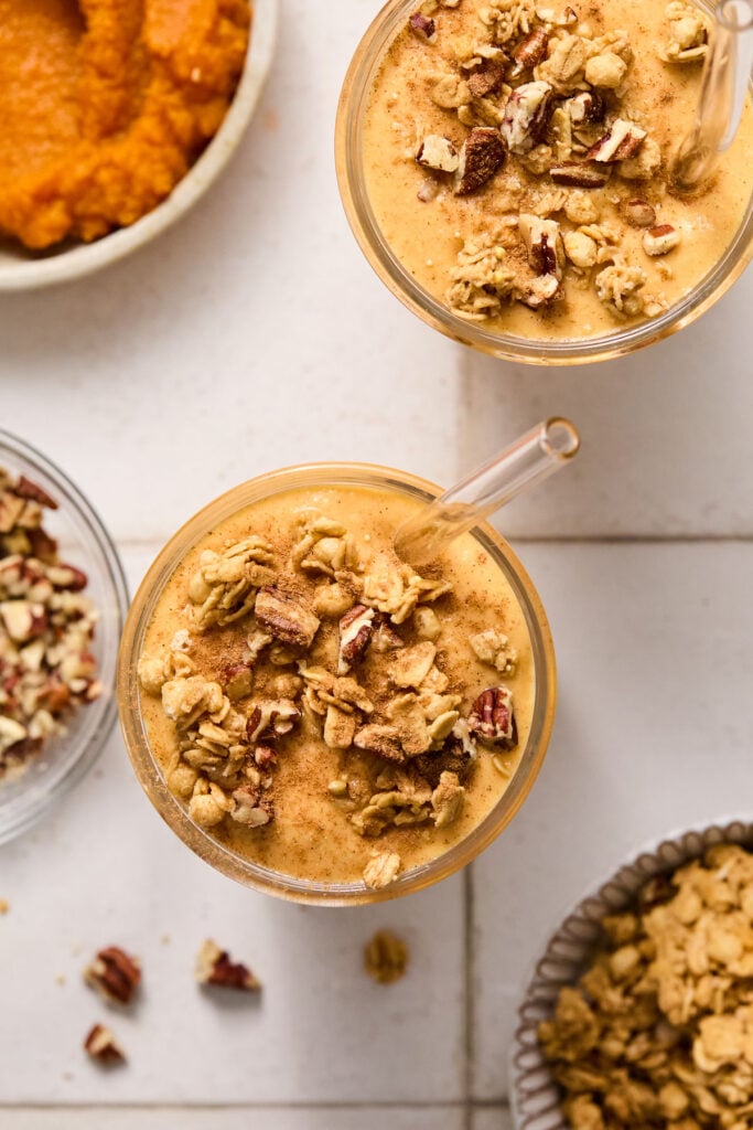 Overhead shot of two pumpkin protein smoothies topped with chopped pecans, granola, and cinnamon in glasses with straws. Around the smoothies are a bowl of granola, a bowl of chopped pecans, and a bowl of pumpkin puree.