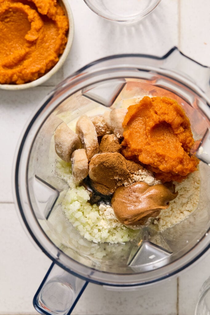 Blender filled with frozen sliced bananas, frozen cauliflower rice, pumpkin puree, vanilla protein powder, vanilla bean paste, almond butter, pumpkin pie spice, salt, and milk. Next to the blender is a bowl of pumpkin puree and an empty glass of milk.
