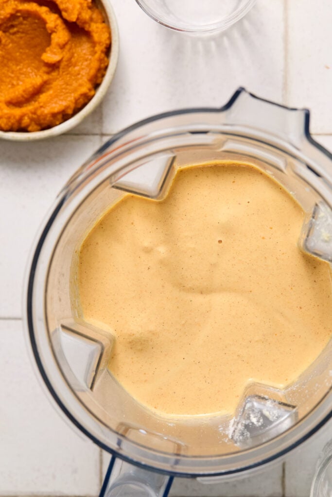 Overhead shot of a creamy orange smoothie in a blender. Next to it is a bowl of pumpkin puree.
