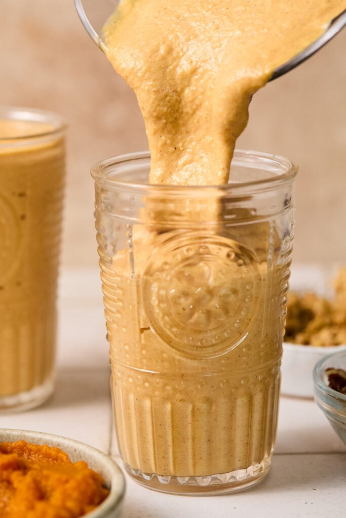 Pumpkin smoothie being poured into a glass from a blender. Around the jar is a bowl of pumpkin puree, another glass of smoothie, and a bowl of granola.