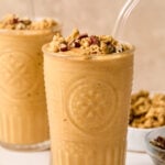 Pumpkin protein shake in a glass topped with chopped pecans and cinnamon, with a straw in it. Around the glass are some chopped pecans, a bowl of pumpkin puree, a bowl of chopped pecans, a bowl of granola, and another glass with a smoothie.