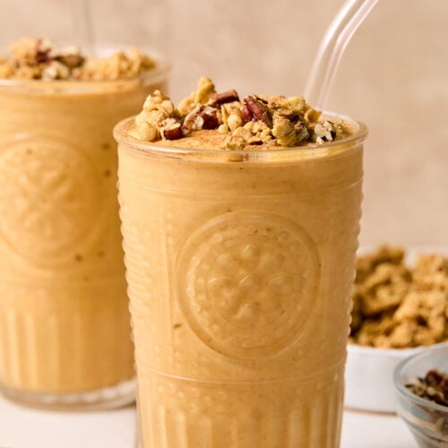 Pumpkin protein shake in a glass topped with chopped pecans and cinnamon, with a straw in it. Around the glass are some chopped pecans, a bowl of pumpkin puree, a bowl of chopped pecans, a bowl of granola, and another glass with a smoothie.